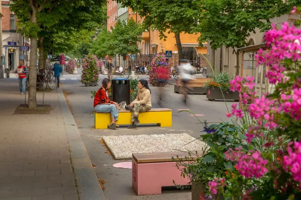 Två personer samtalar på färgglada bänkar omgivna av blommor på en bilfri sommargata.