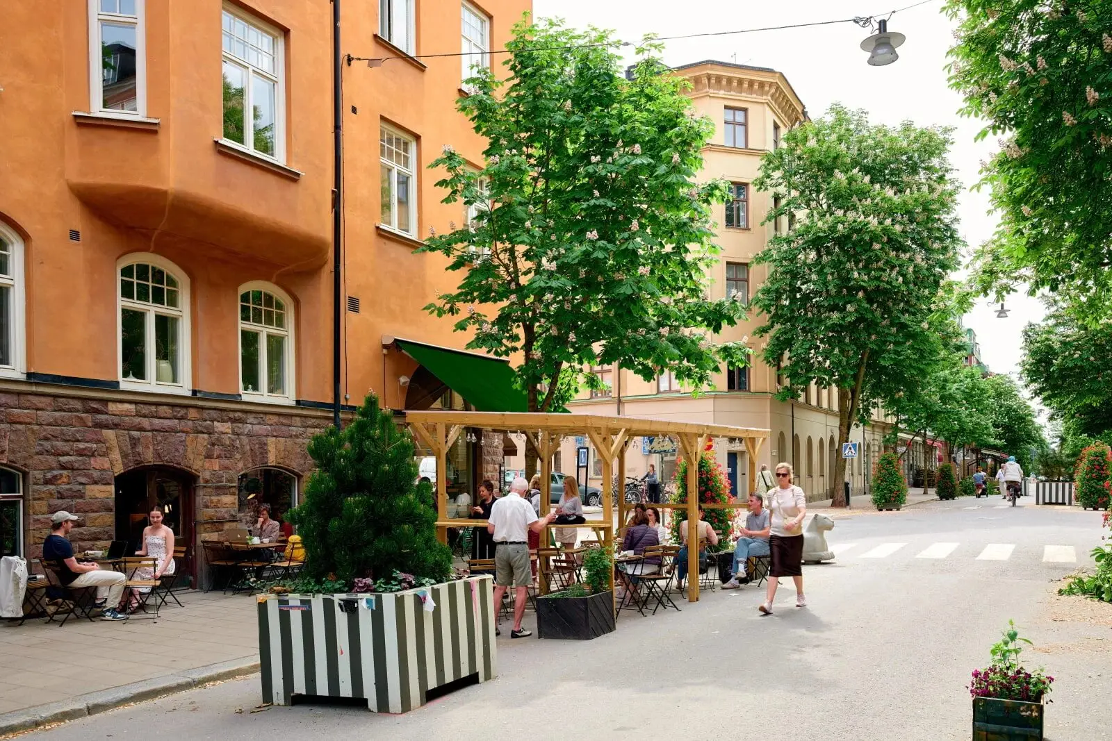 Uteservering under blommande kastanjeträd på en bilfri sommargata i Stockholm, människor sitter vid bord ute på gatan.