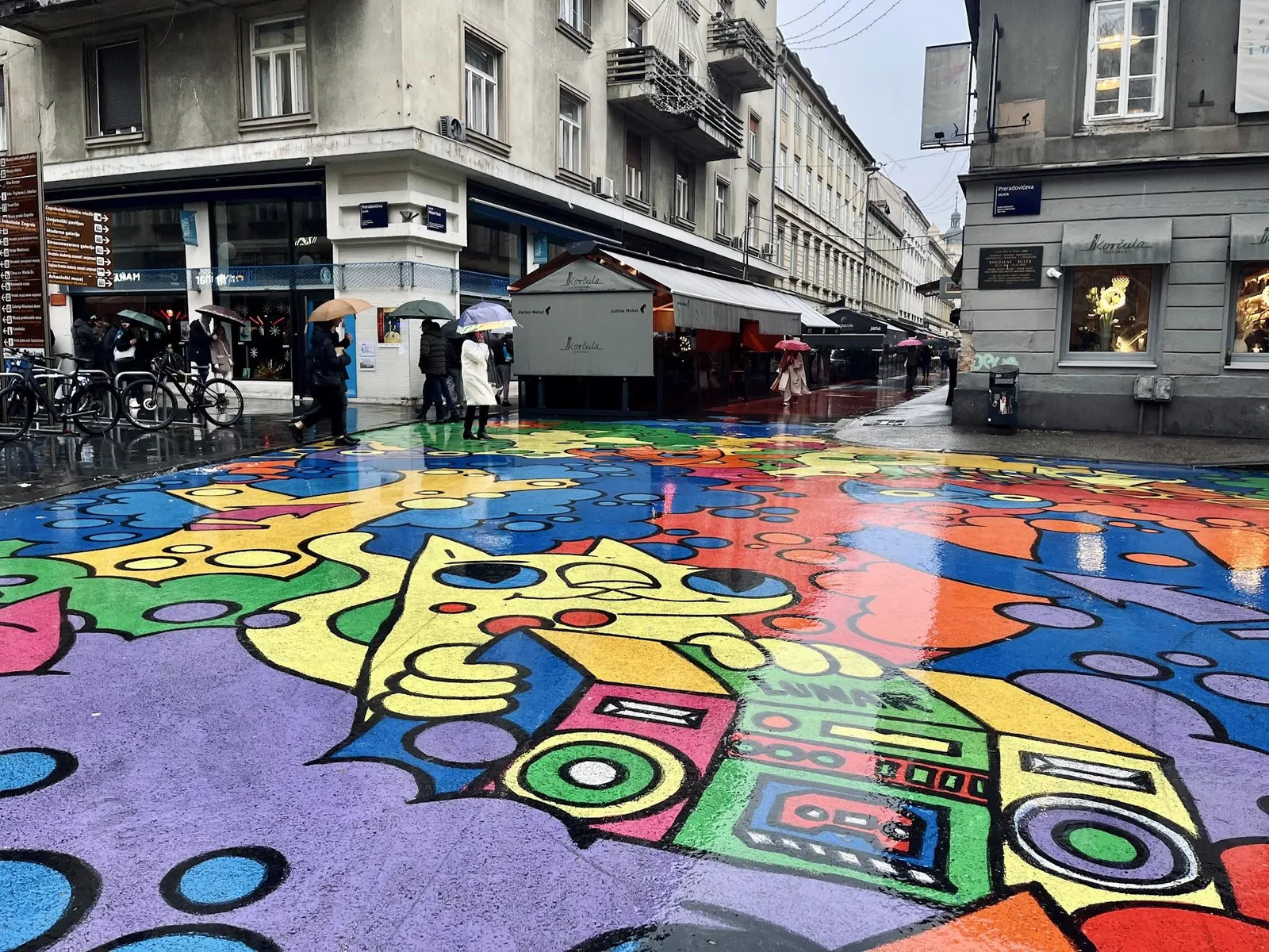 Ett gathörn i centrala Zagreb där hela körbanan är täckt av en färgstark muralmålning, med människor som går under paraplyer.