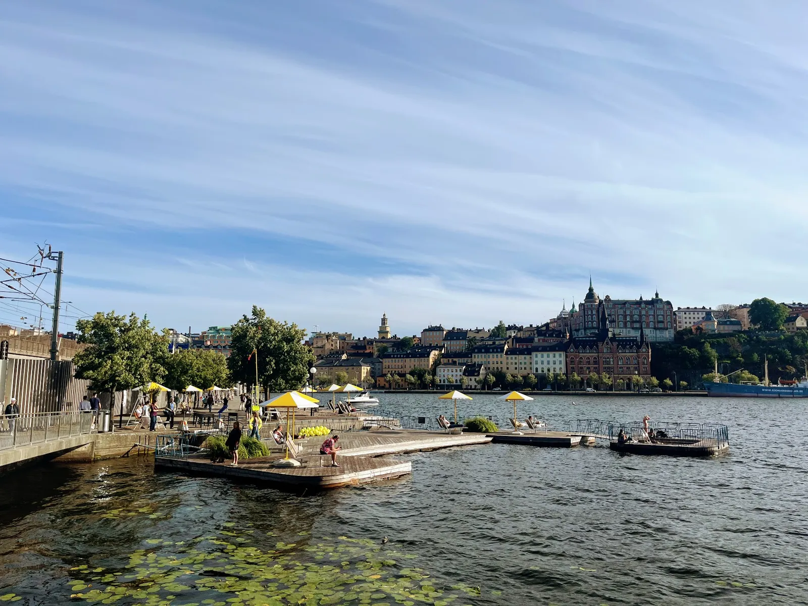 Munkbrohamnen: badbryggor i trä med gula parasoller som flyter på vattnet, med Södermalms siluett bakom.