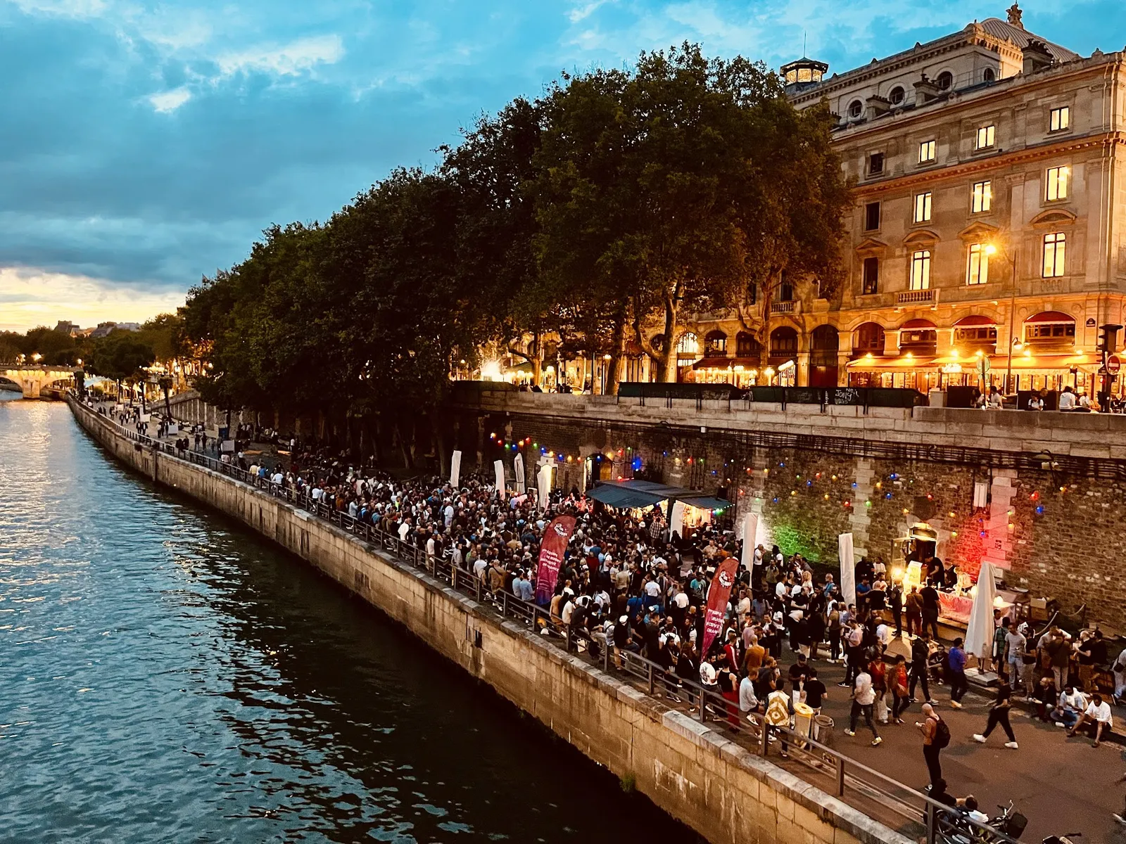 Paris Plages i skymningen: folksamlingar längs Seines kaj under träd och ljusslingor, med kaféer som lyser i bakgrunden.